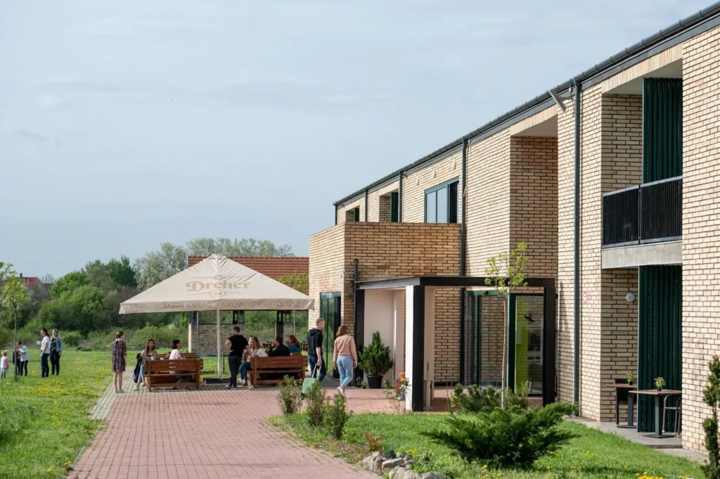 Modern, téglaburkolatú szállodaépület előtt napernyő alatt ülő vendégek egy zöld réten.