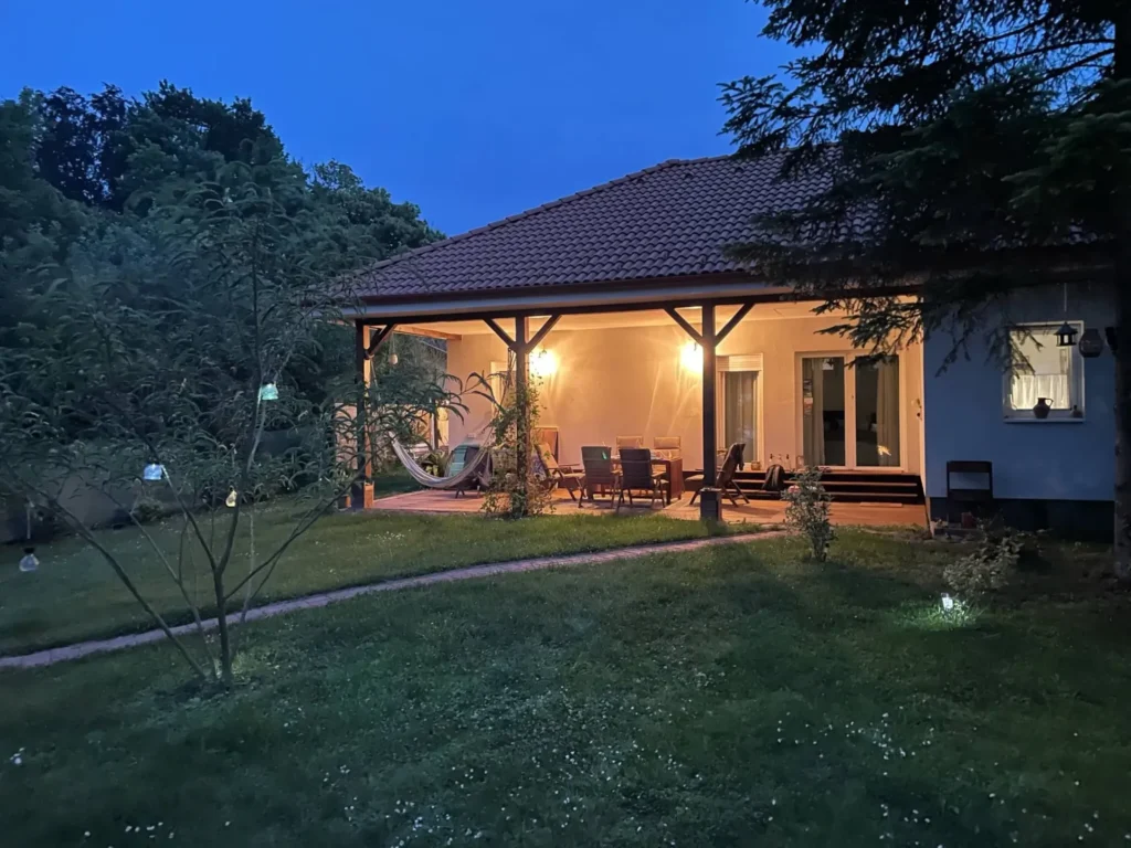 Esti hangulatú, kivilágított teraszos vendégház zöld kerttel és fákkal körülvéve.