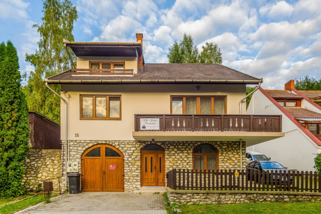 Kőalapzatú, erkélyes családi ház fa kapukkal és kerítéssel, felhős égbolt alatt.